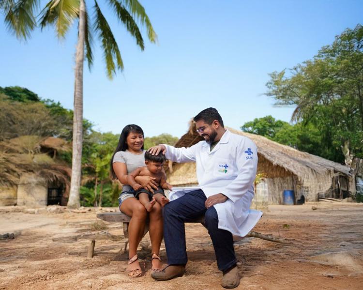 Homem de jaleco branco olha com carinho para criança indígena acompanhada de sua mãe. Eles estão sentados ao ar livre