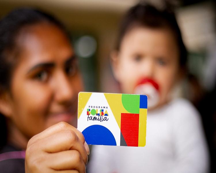 Cartão do Bolsa Família em primeiro plano é mostrado por mulher com bebê no colo