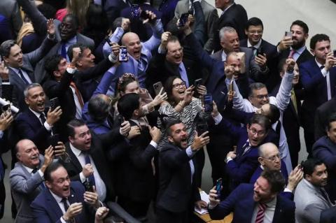 Homens de terno e uma mulher olham para o alto com braços erguidos e apontando celulares