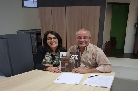 Uma mulher e um homem sentados a uma mesa de trabalho mostram um livro. Eles sorriem