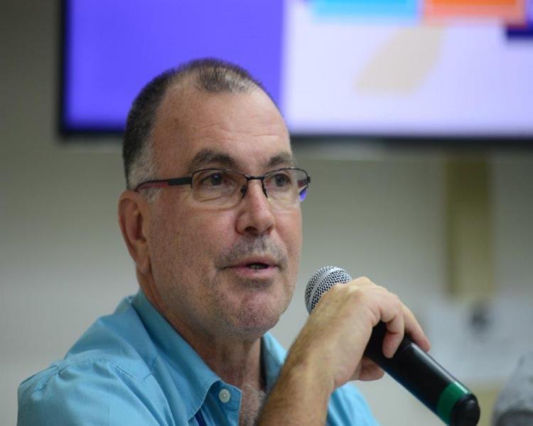 Homem em primeiro plano, de óculos e camisa azul, ao microfone