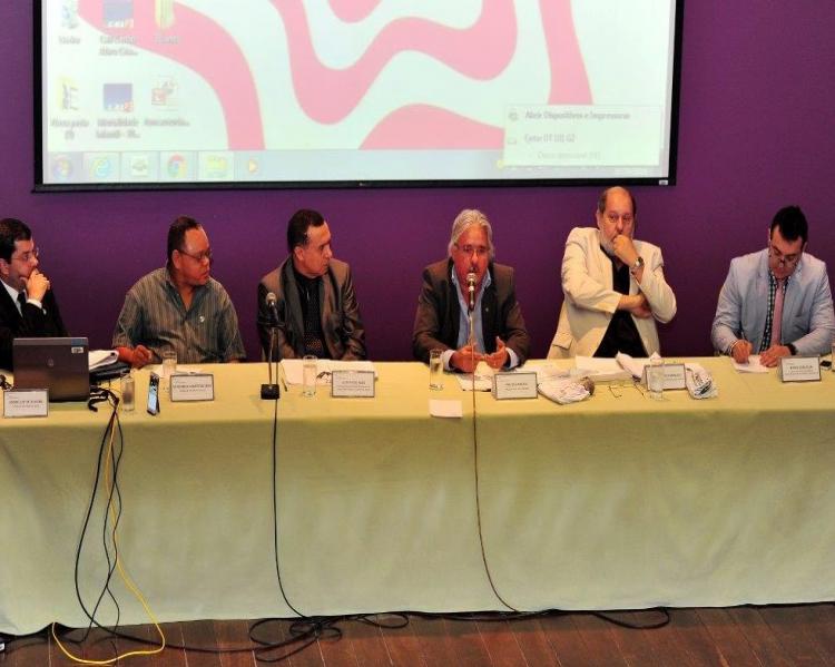 Seis homens em uma mesa de debates, um deles ao microfone