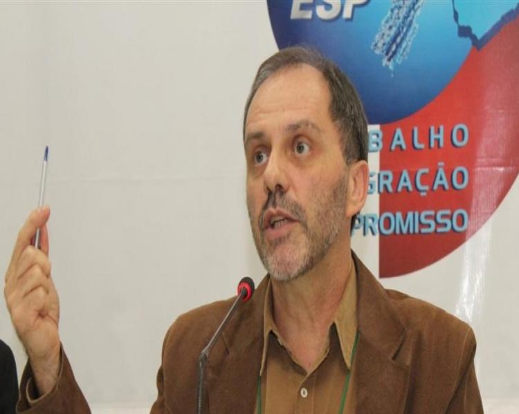 Homem de paletó e camisa sem gravata, sentado à mesa de debates fala ao microfone e segura uma caneta em uma das mãos