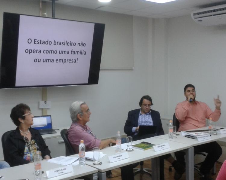 Uma mulher e três homens em mesa de debates, tendo o da ponta direita falando ao microfone, assistido pelos demais; atrás e acima deles, um telão para apresentaões, onde se lê: ''o Estado brasileiro não opera como uma família ou uma empresa!