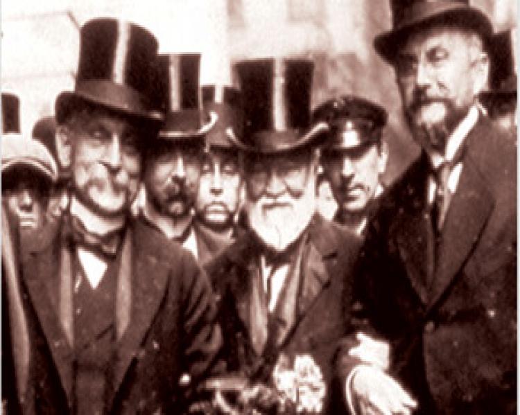 Homens em trajes antigos, com cartolas e fraques, em primeiro plano. Foto em preto e branco