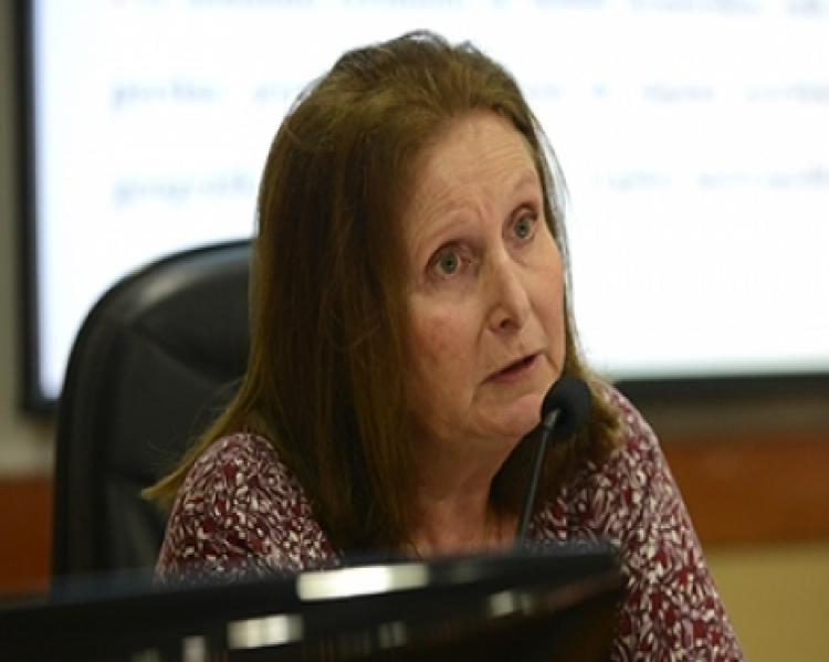 Mulher de cabelos claros e ongos de perfil em close, sentada em uma mesa de debates