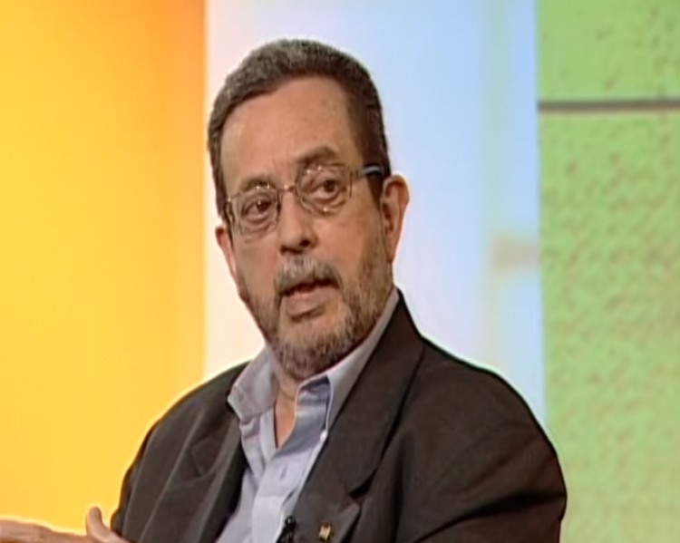 Homem de barba e óculos, vestindo paletó escuro, em fundo colorido