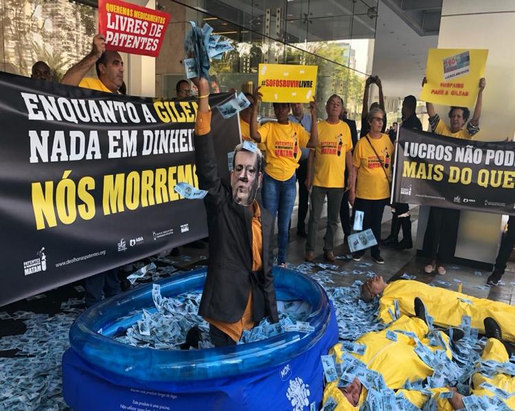 Homem dentro de uma piscina de plástico, com notas de dinheiro a sua volta e pelo chão, onde há pessoas deitadas; ao fundo, ativistas com cartazes contra patentes e com os dizeres 'Enquanto a Gilead nada em dinheiro, nós morremos'