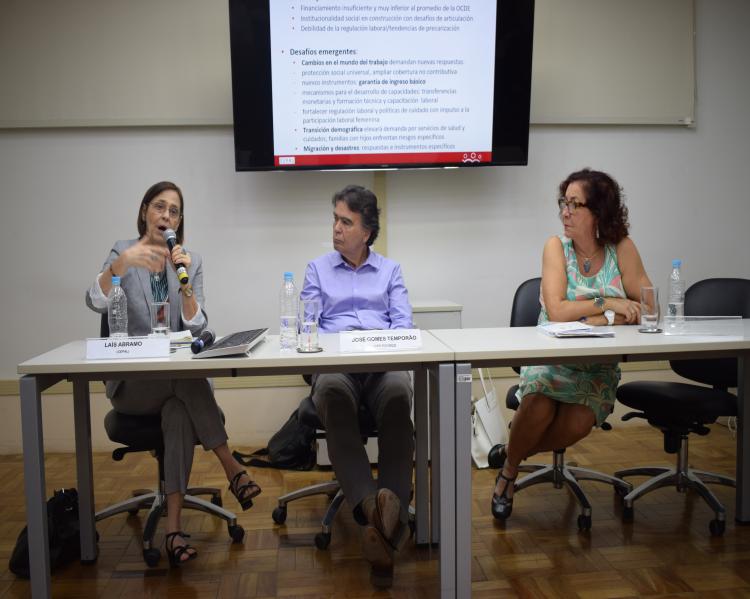 Três pessoas, duas mulheres e um homem ao centro, em uma mesa de debates, tendo-se ao fundo  um telão 