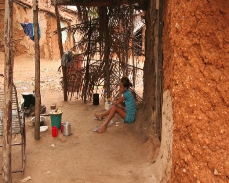 Criança ao longe sentada no chão de terra, na porta de casa