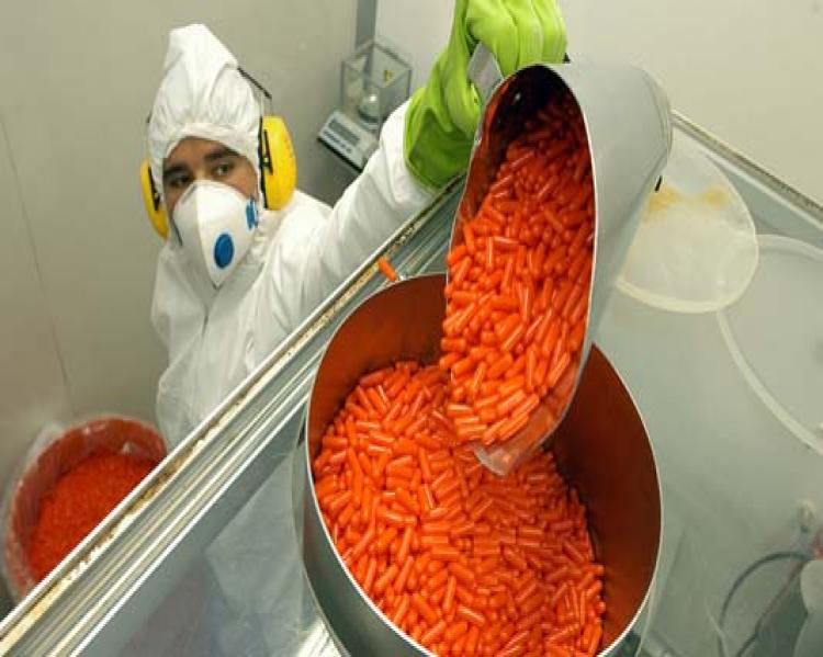 Recipiente com comprimidos de cor laranja manuesado por pessoa, ao fundo, com jaleco, máscara e touca