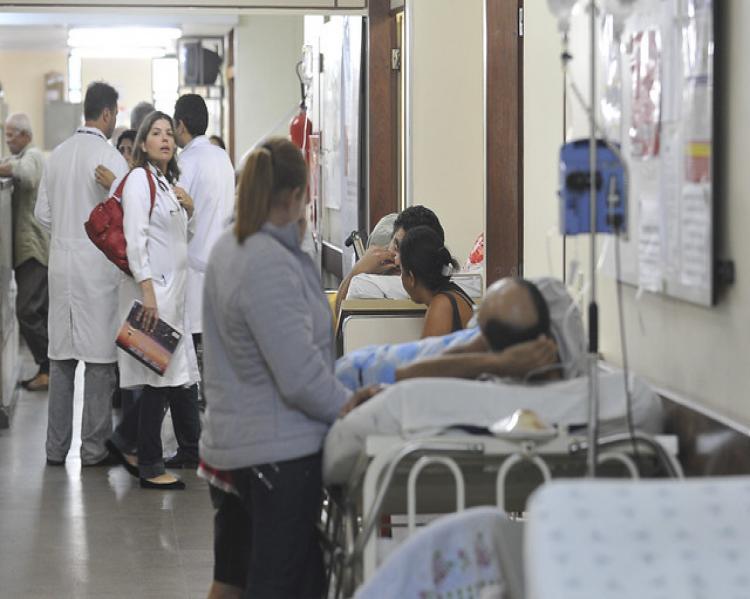 Corredor de hospital com macas enfileiradas, pessoas deitadas, uma mulher na cabeceira de uma das macas