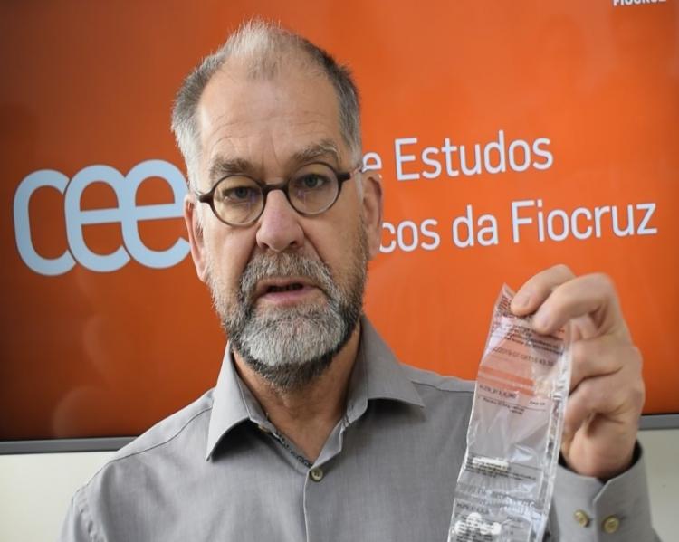 Homem de barba grisalha e óculos mostra uma tira plástica contendo comprimidos, em fundo laranja mostrando a logomarca do Centro de Estudos Estratégicos da Fiocruz