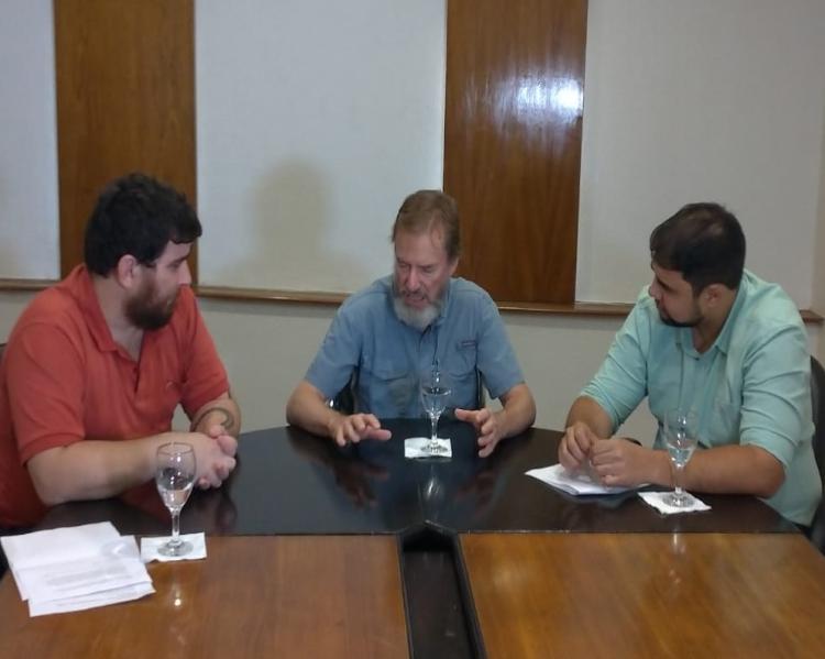 Três homens sentados a uma mesa; dois deles, nas pontas, prestam atenção à fala do que está no centro