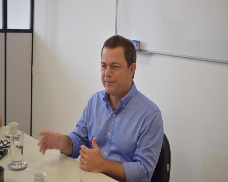 Homem de camisa social azul, sentado a uma mesa, fala e gesticula