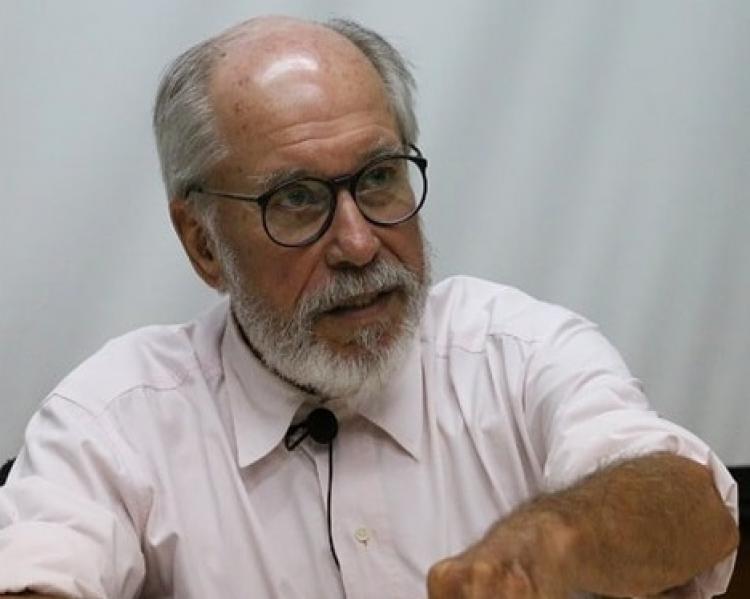 Homem grisalho de barba e óculos, de camisa clara, com microfone preso à lapela, fala e gesticula, sentado a uma mesa