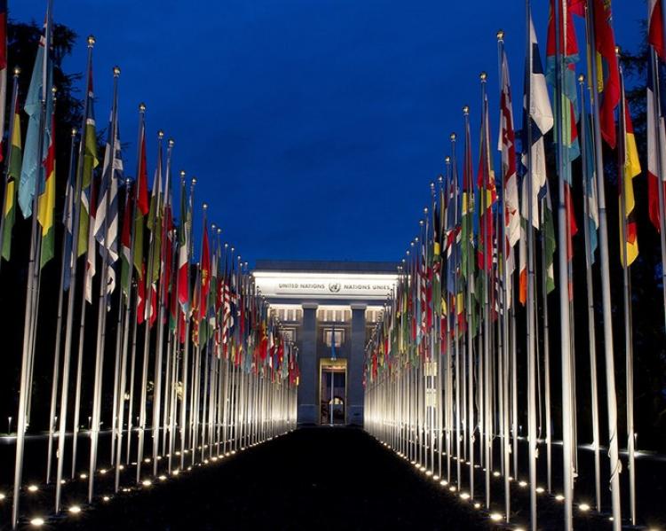 Duas fileiras de bandeiras de países, com a sede das Nações Unidas ao fundo, sob céu azul