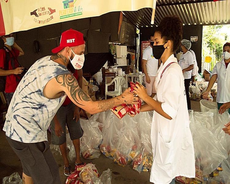 Homem de camiseta, boné vermelho e máscara recebe alimentos das mãos de uma mulher de jaleco e máscara,em meio a sacos com cestas básicas