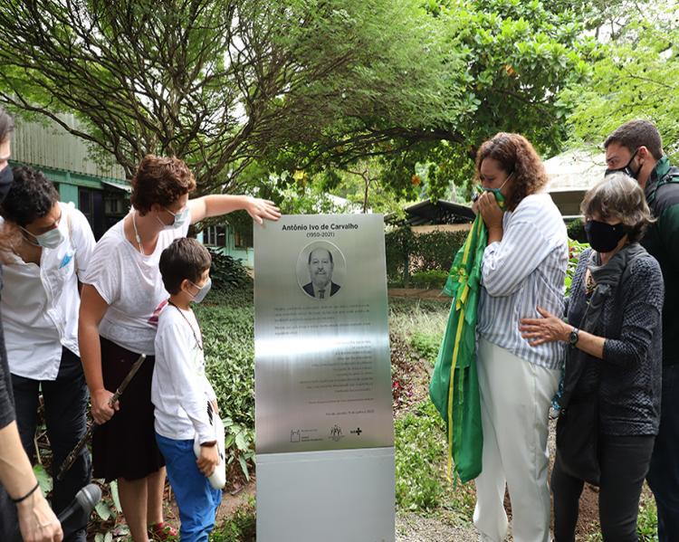 Pessoas ladeando uma placa onde se vê um homem sorridente e um texto