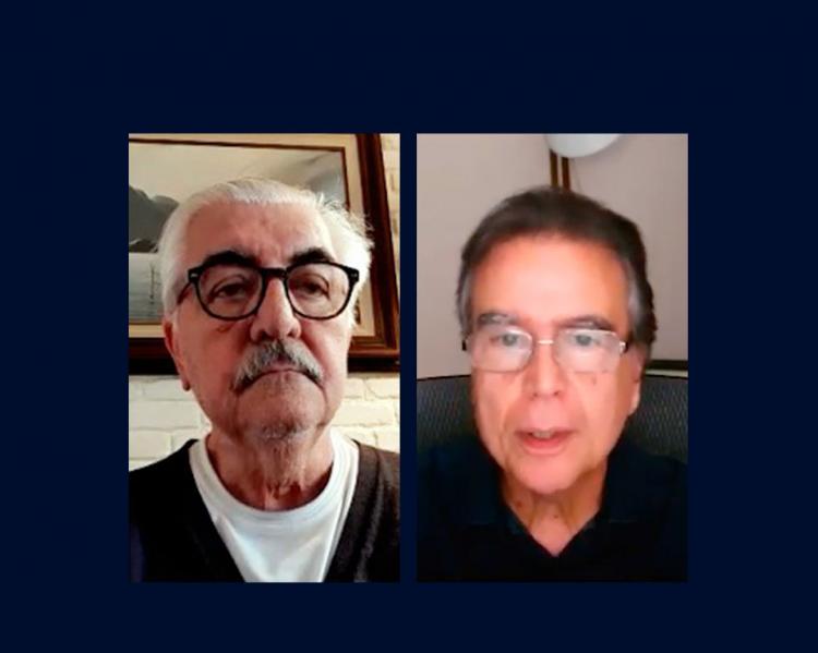 Dois homens em close, lado a lado, um grisalho, de bigode e de óculos de aro escuro, outro de cabelos escurso e óculos de aro prateado