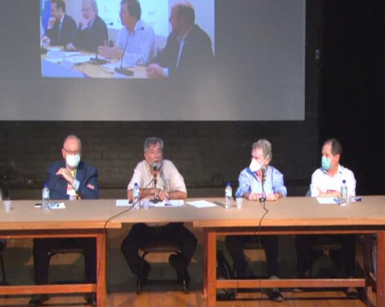 Quatro homens sentados a uma mesa longa, um deles fazendo uma apresentação