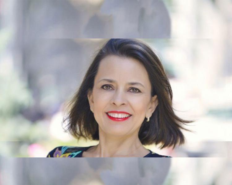 Rosto de mulher de cabelos pretos, esvoaçantes, em local aberto. Ela usa batom avermelhado e sorri levemente