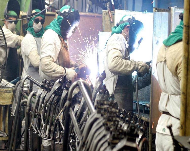 Homens com proteção de acrílico transparente no rosto manuseiam equipamentos em uma fábrica