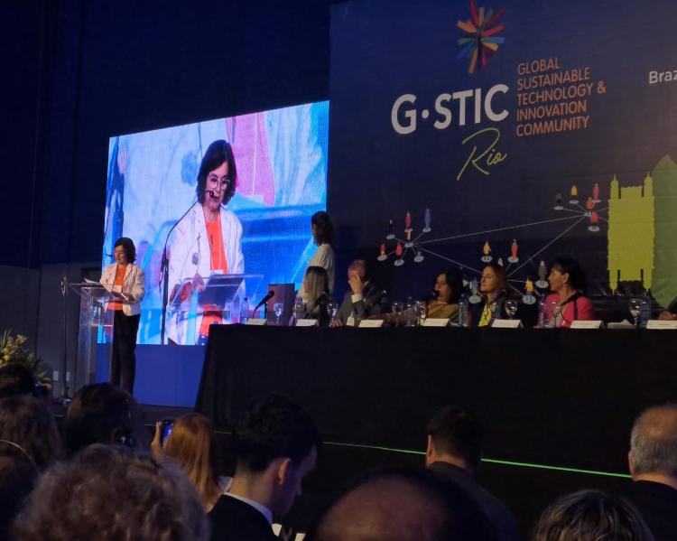 Mulher de pé ao fundo, fala em um púlpito observada por pessoas sentadas a uma mesa num palco