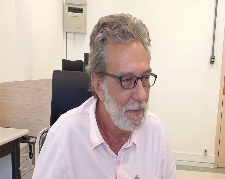 Homem grisalho de óculos, vestindo camisa rosa clara, está sentado em um ambiente de trabalho