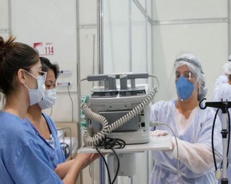 Equipe de trabalhadoras de saúde algumas de jalecos azuis, outras, de branco, usando máscara, estão em volta de uma máquina em uma sala hospitalar
