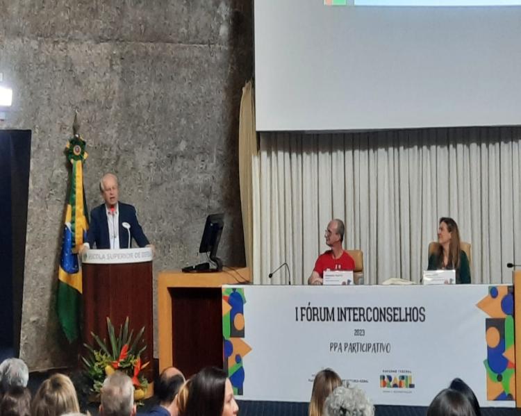 Homem de terno azul fala em um púlpito, observado por um homem e uma mulher sentados em uma mesa de evento