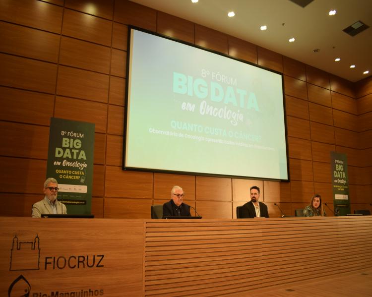 Três homens e uma mulher em uma mesa de debates tendo atrás um telão onde se lê Oitavo Fórum Big Data