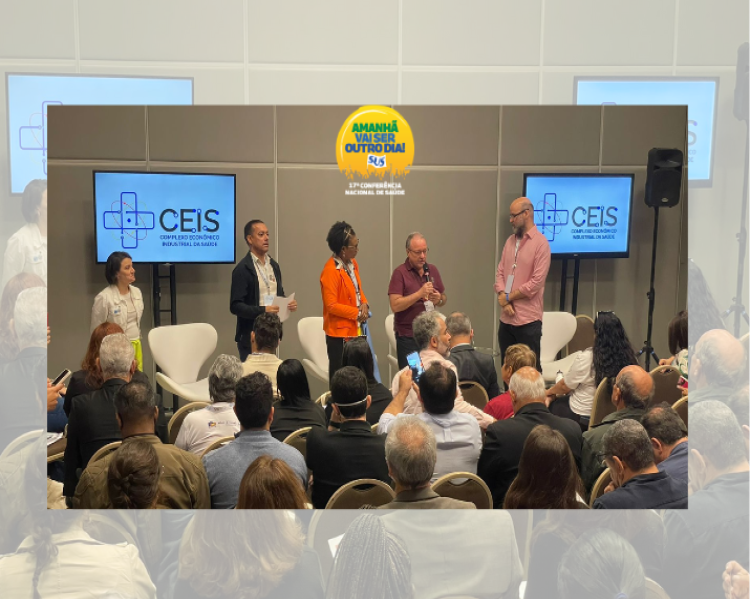 Dois homens e uma mulher de pé, à frente de uma plateia. O homem ao centro fala aos demais presentes