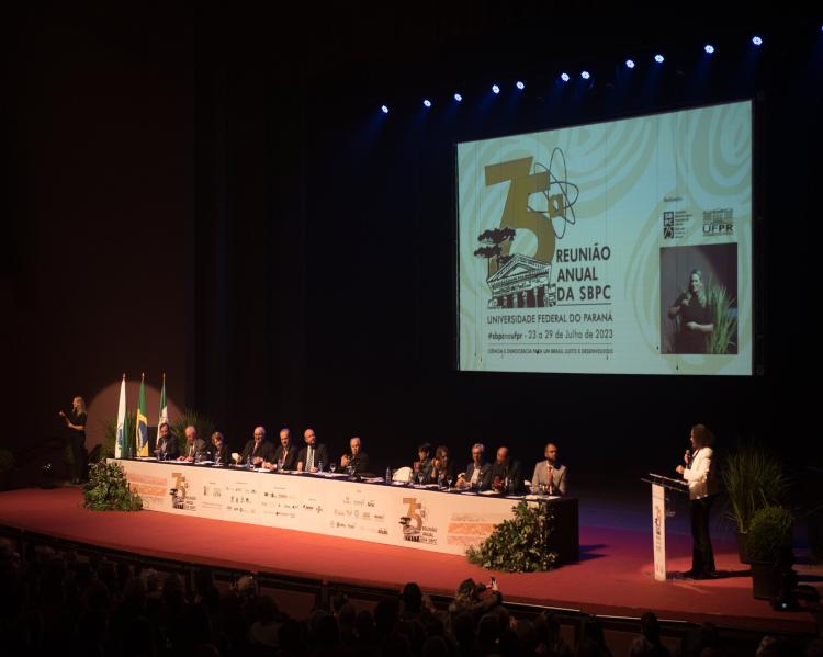 Longa mesa de debates em um palco, onde homens e mulheres estão sentados tendo atrás um telão com a mara da 75ª reunião anual da sbpc