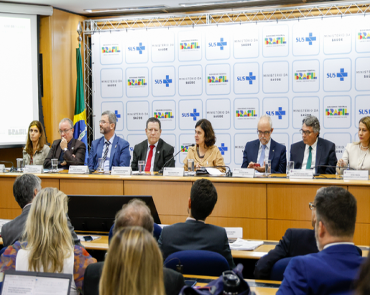 Mulheres e homens sentados a uma mesa de debates tendo ao fundo logos do SUS e do Governo Federal