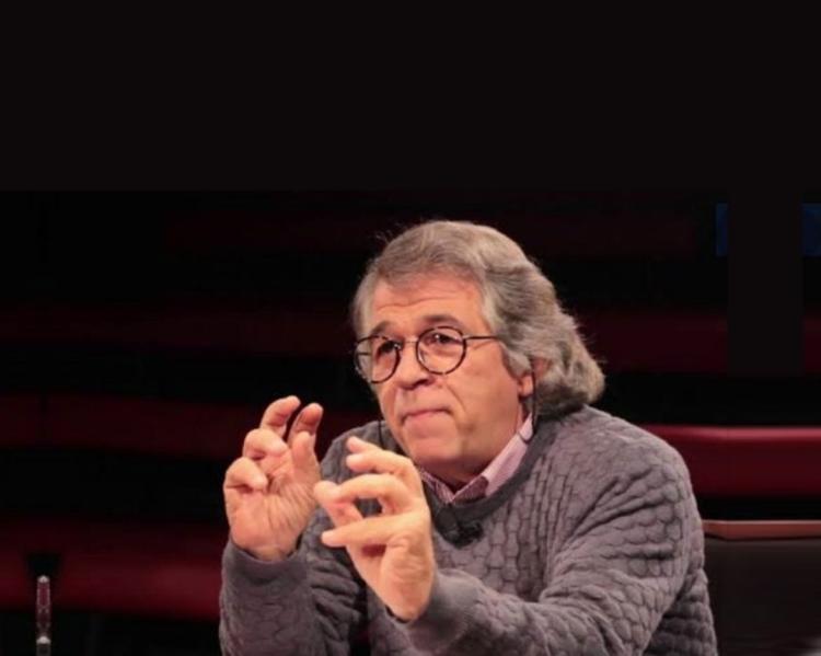 Homem de óculos e gesticula sentado a uma mesa