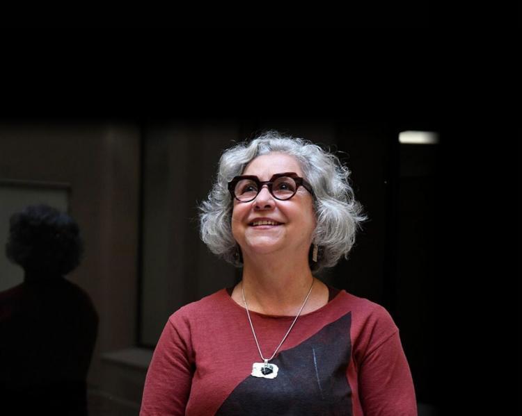 Mulher de cabelos grisalhos, óculos de aro preto e blusa vermelha sorri olhando para o alto