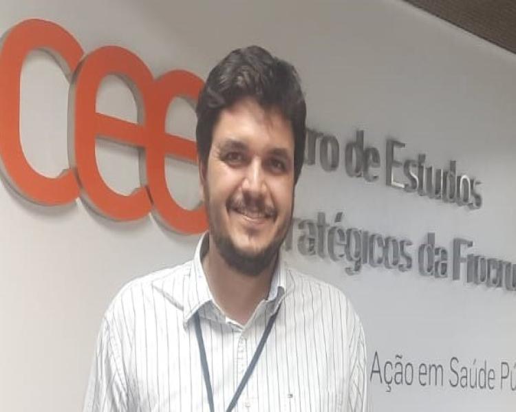 Homem de barba e cabelos castanhos, de pé, com as mãos no bolso da calça e camisa social branca, à frente de uma parede branca com a logomarca do CEE