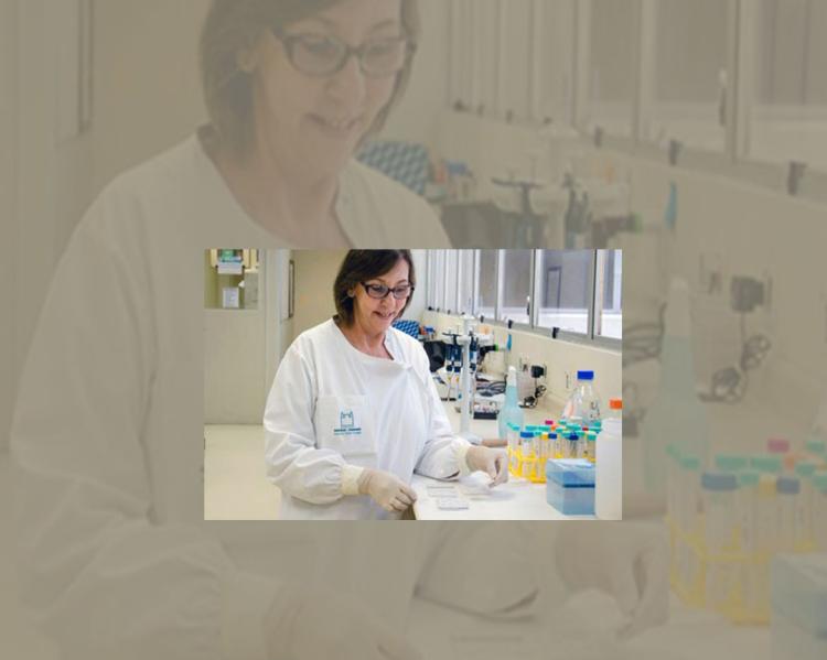 Mulher de óculos e jaleco manipula material sobre bancada de laboratório