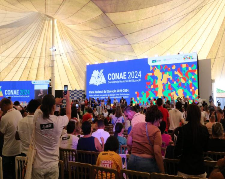 Plenária lotada de homens e mulheres tendo ao fundo o palco com painel com a logomarca da Conae 2024