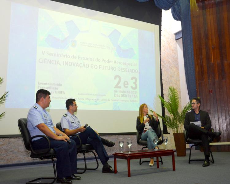 Três homens e uma mulher sentados em cadeiras sobre um palco; a mulher fala ao microfone