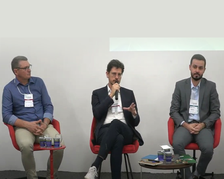 Três homens sentados em suas cadeiras lado a lado; o do meio está falando ao microfone, observado pelos demais