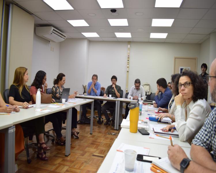 Mulheres e homens em uma extensa mesa de debates, em forma de U, um deles, ao fundo, fala ao microfone