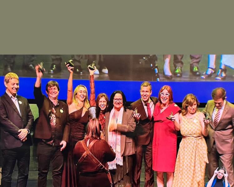 Homens e mulheres levantam estatuetas de premiação em um palco; estão felizes