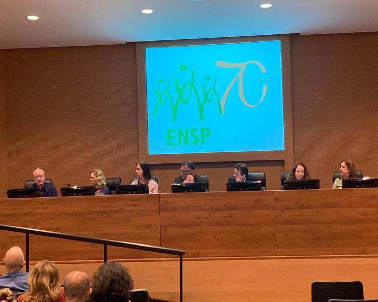 homens e mulheres em uma bancada no palco de um auditório, com telão atrás. Um deles fala para a plateia