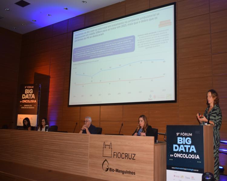 Homens e mulheres em uma mesa de debates sobre um palco. Em primeiro plano uma mulher fala em um púlpito. Ao fundo um telão com informações sobre a palestr