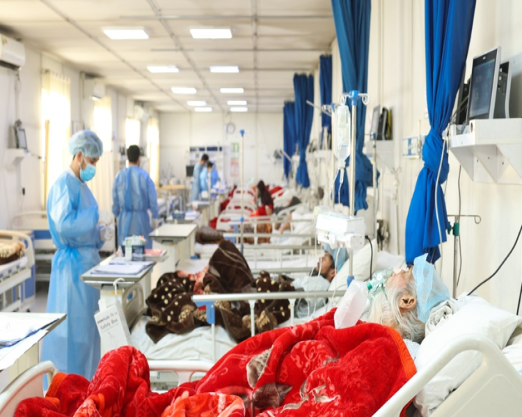 Enfermaria hospitalar, com macas enfileiradas e médicos com jalecos azuis em pé próximos aos leitos