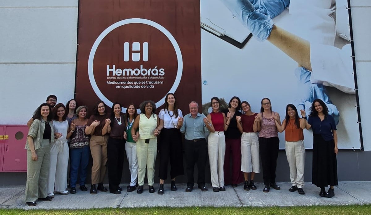 Homens e mulheres estão em pé tendo ao fundo a logomarca da Hemobrás