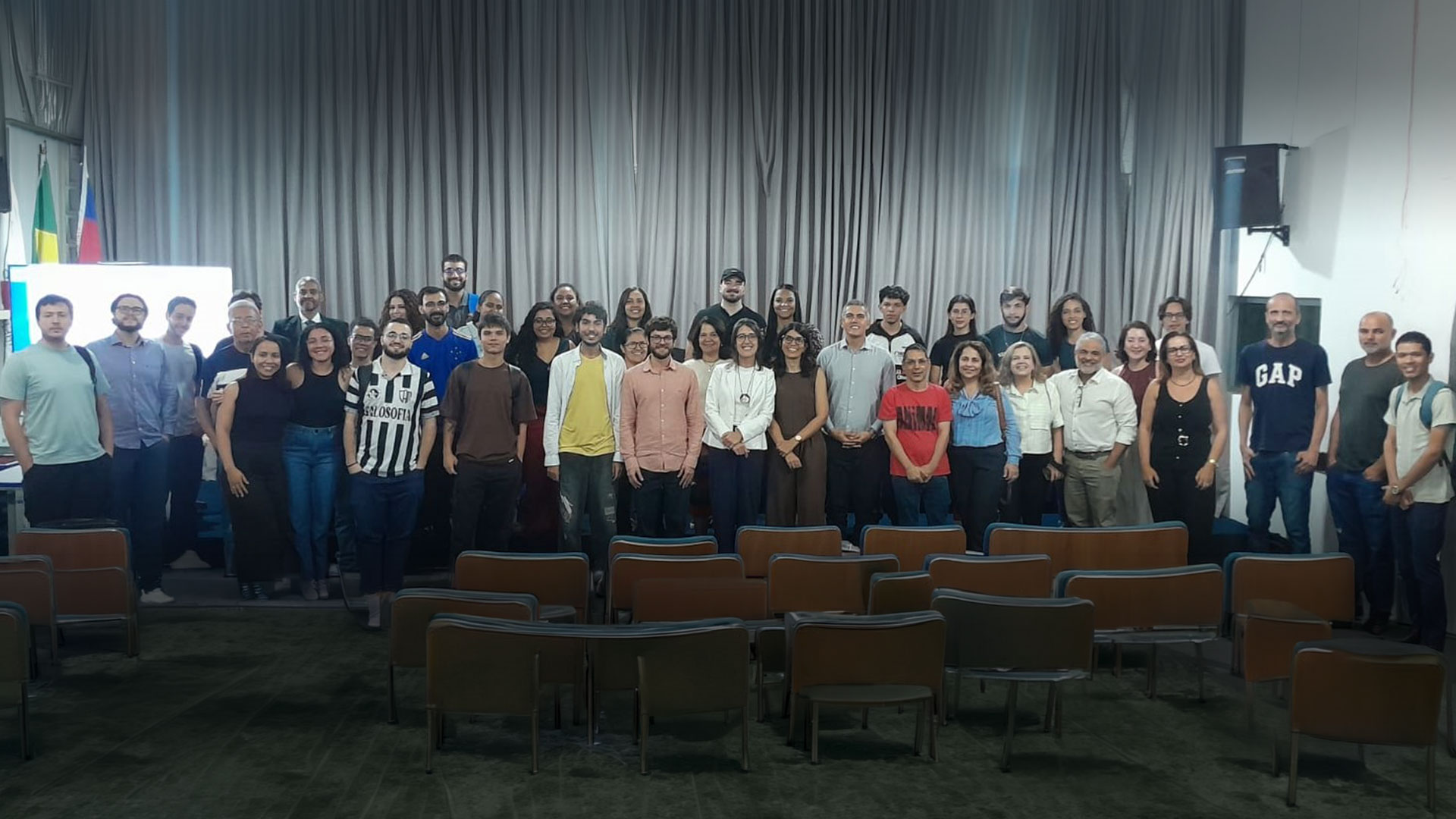 Homens e mulheres, jovens e adultos, estudantes e professores, estão de pé e sorriem, após aula inaugural