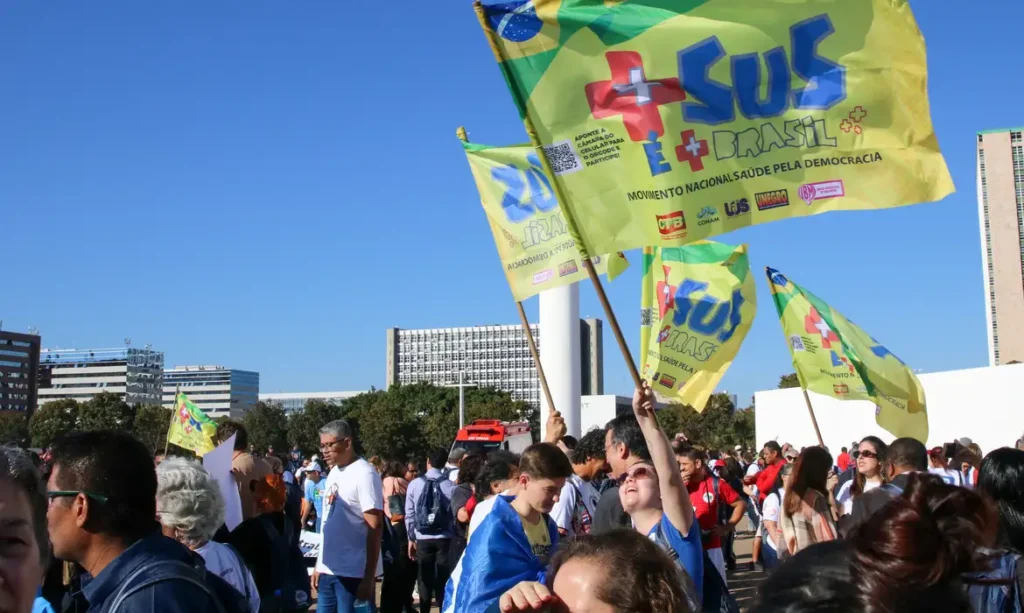 Bandeiras de fundo amarelo com a logomarca do SUS e e o texto Movimento Nacional Saúde pela Democracia tremulam sob o céu azul, levada por pessoas reunidas em uma manifestação